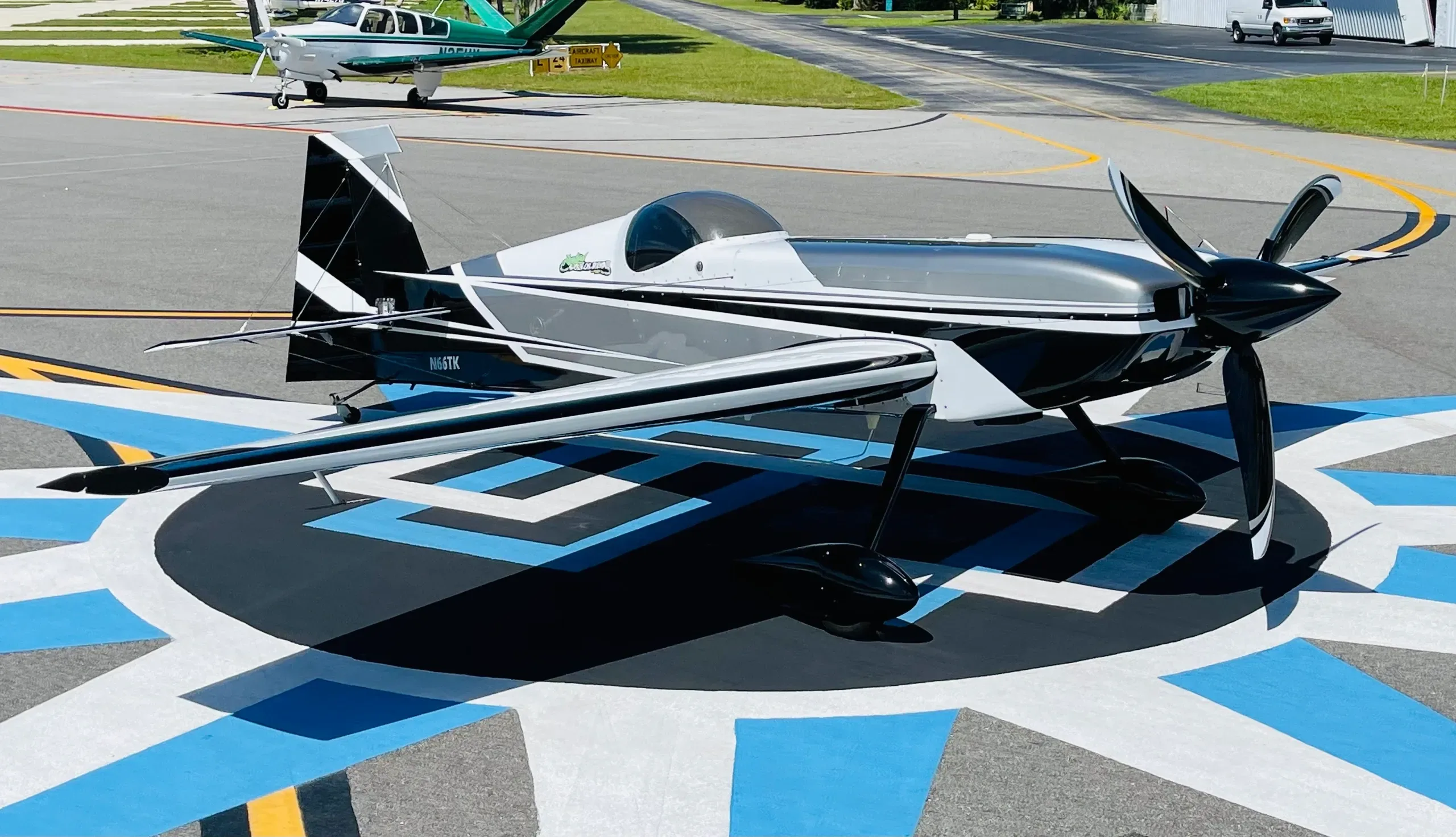A black and white aerobatic aircraft (N66TK) with a distinctive paint scheme parked on a blue and black patterned tarmac.