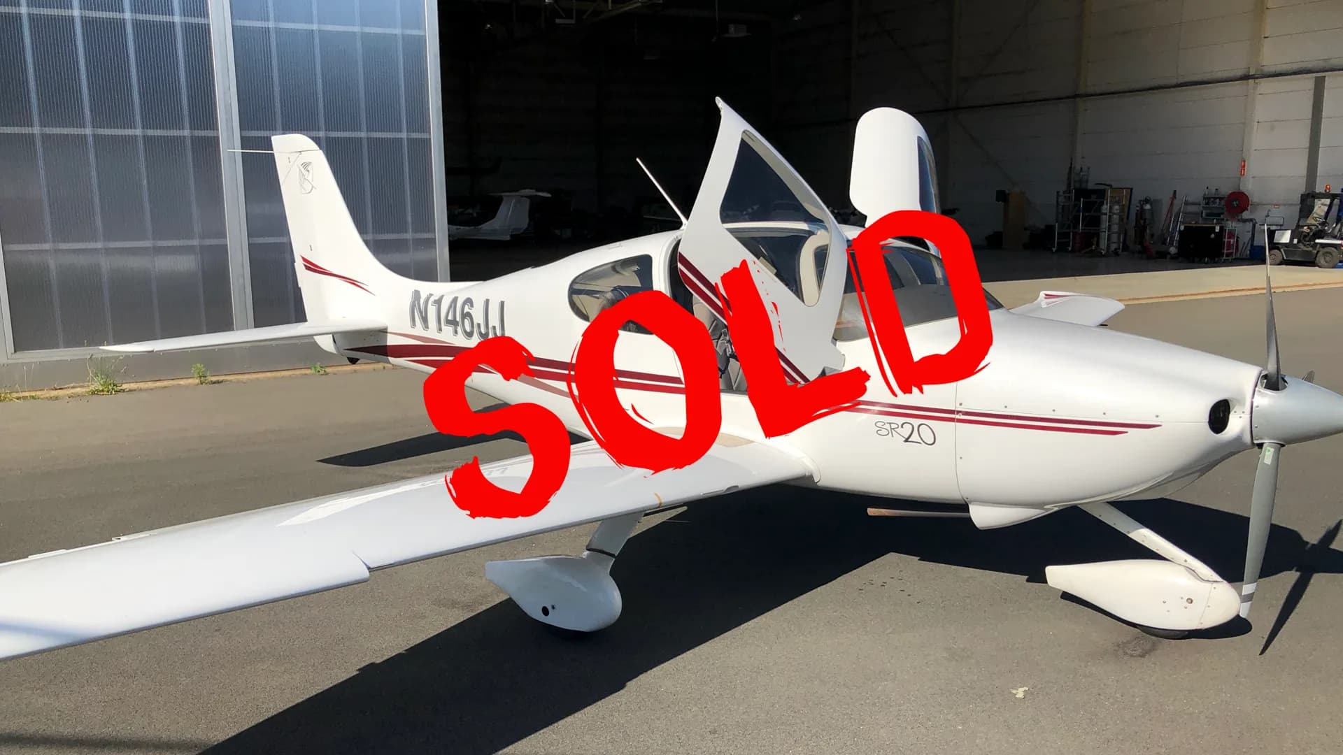 A white Cirrus SR20 aircraft with a red stripe, parked on an apron in front of a hangar, with its doors open.