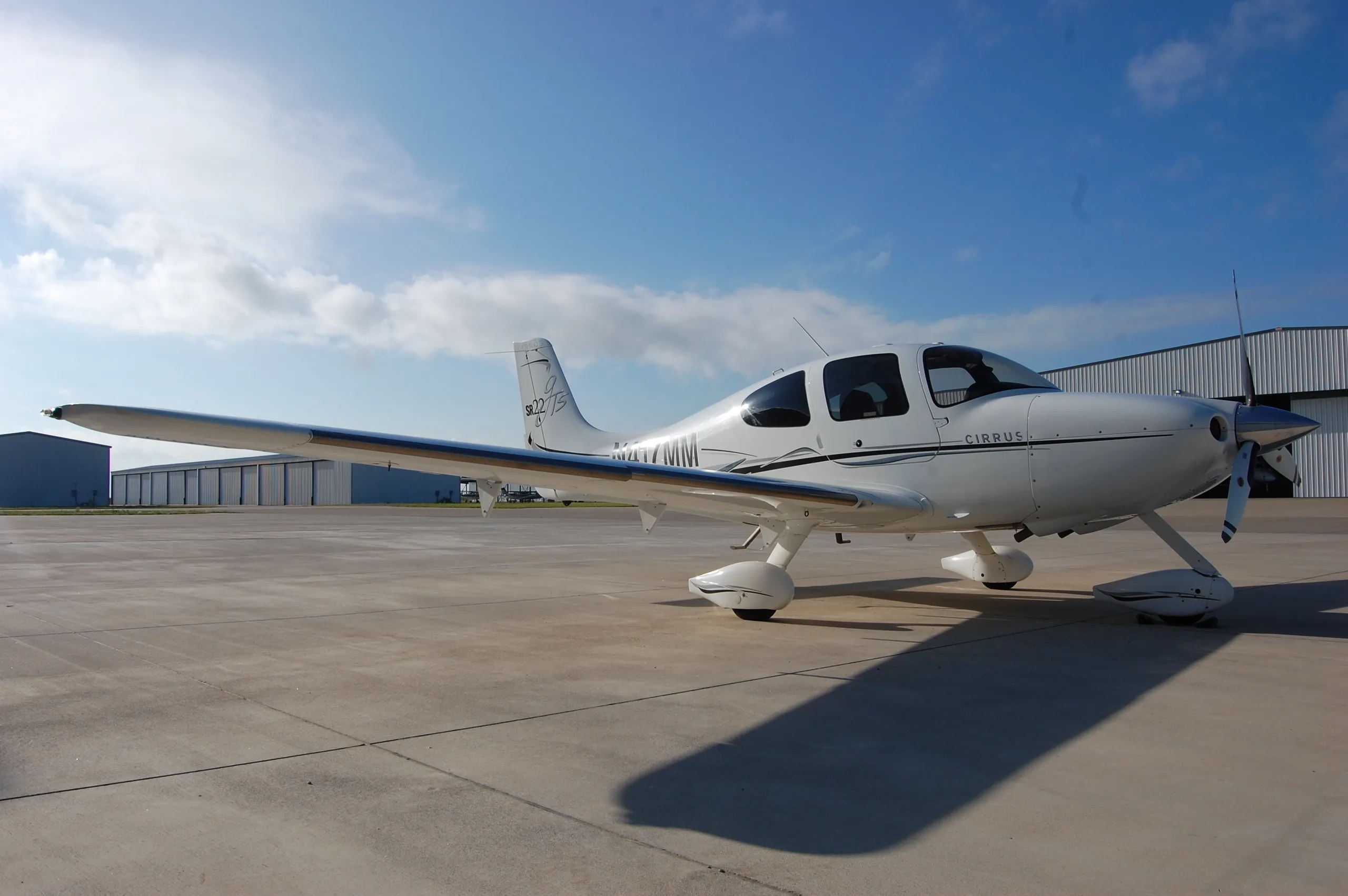 A white Cirrus SR22 aircraft with black and grey stripes parked on an airfield with hangars in the background.