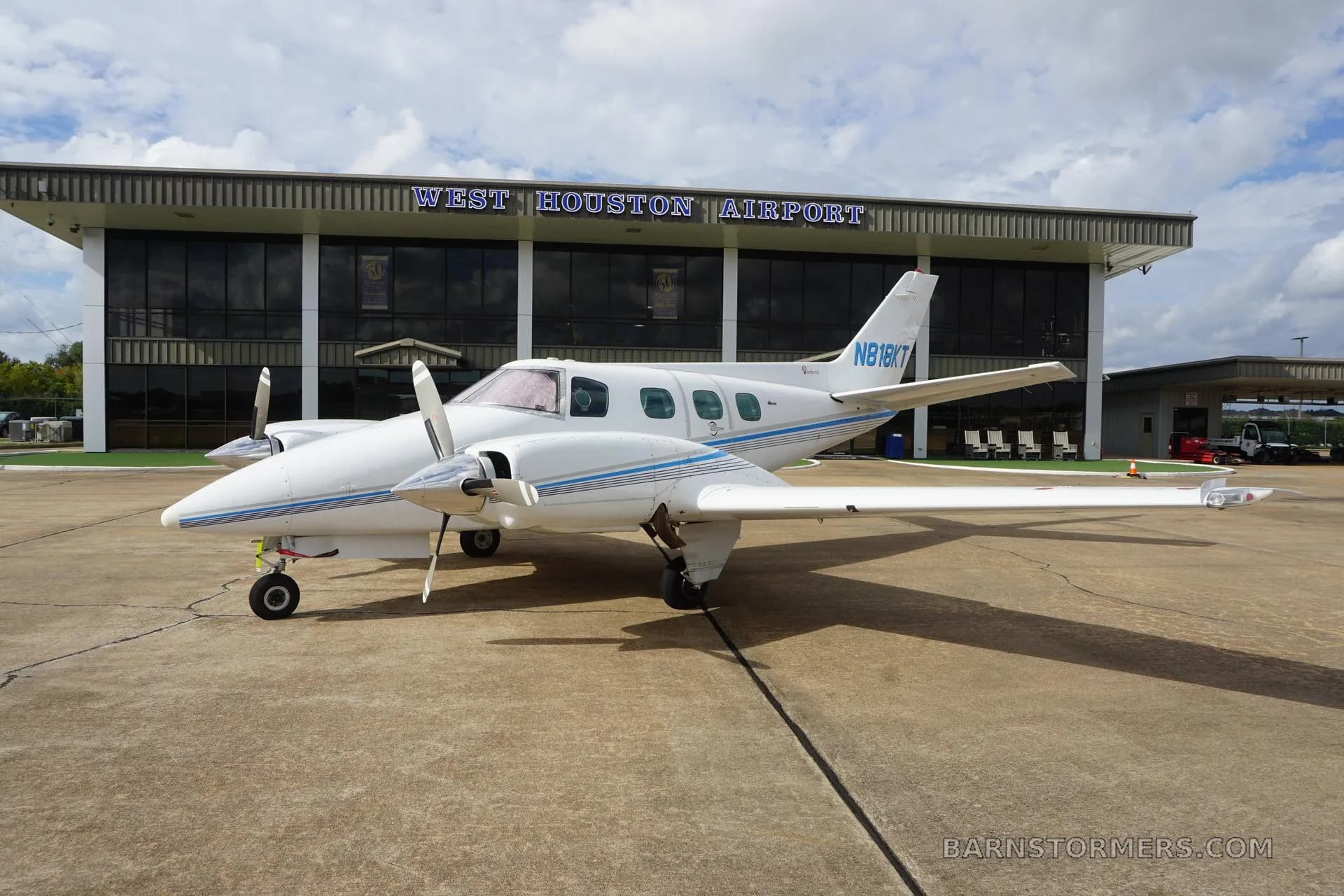 A white twin-engine propeller aircraft with blue stripes (N818KT) is parked on the tarmac in front of the West Houston Airport building.
