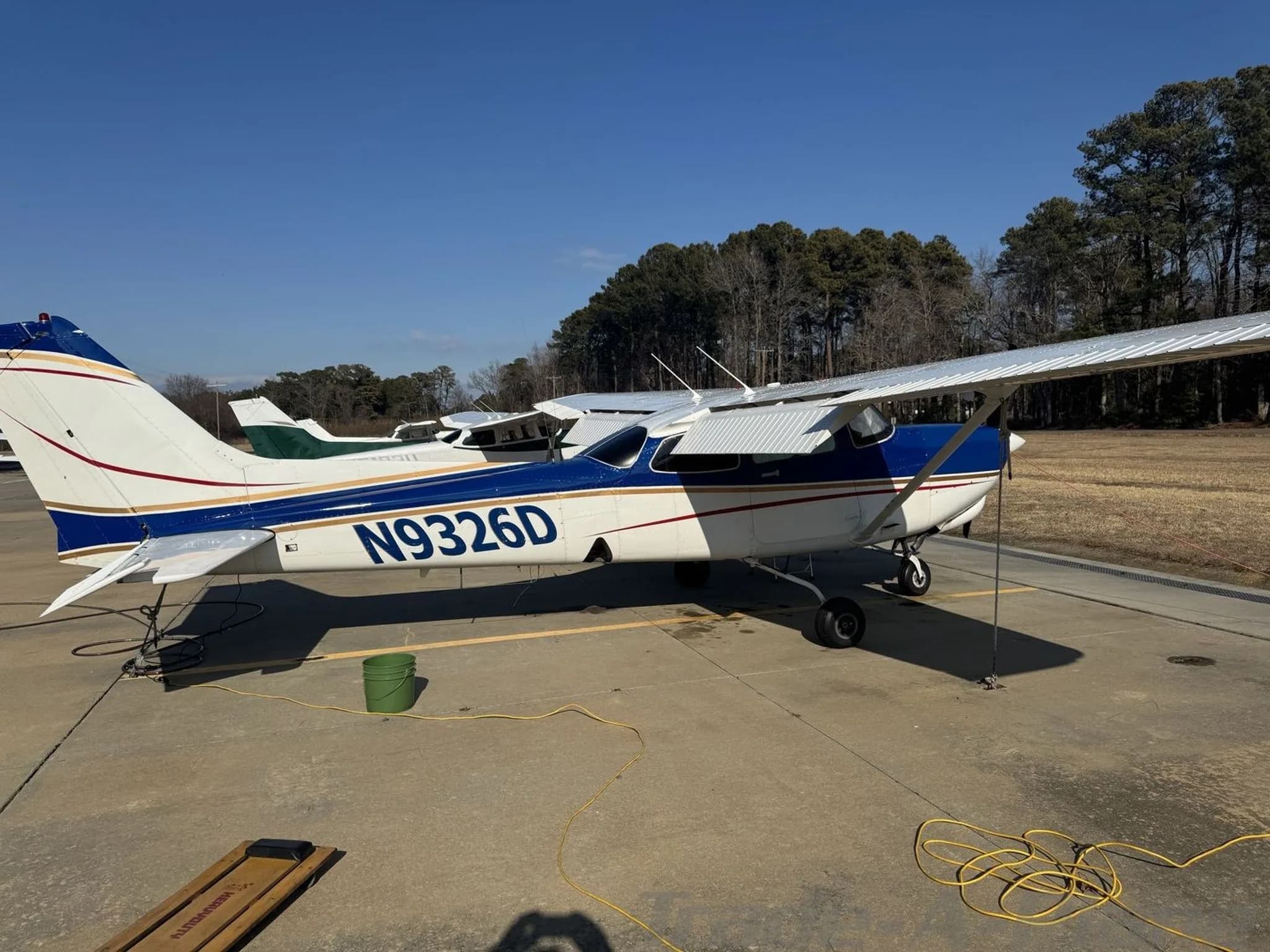 A blue and white Cessna 182 Skylane with registration N9326D parked on an airfield under a clear sky.