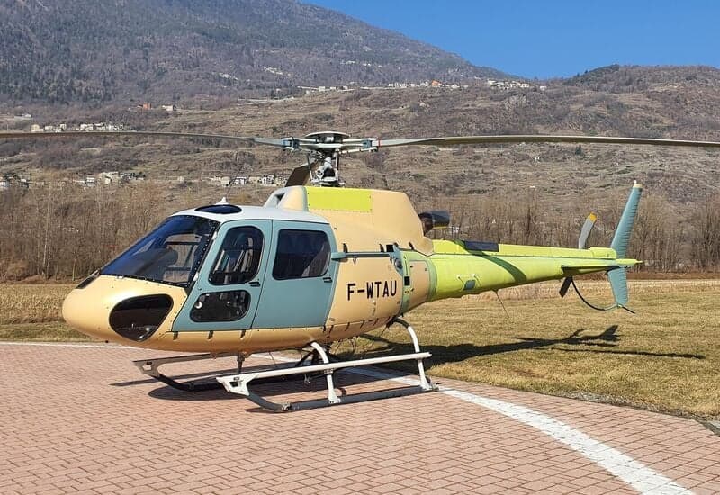 A Eurocopter AS350 Ecureuil helicopter is parked on a helipad with mountains in the background.