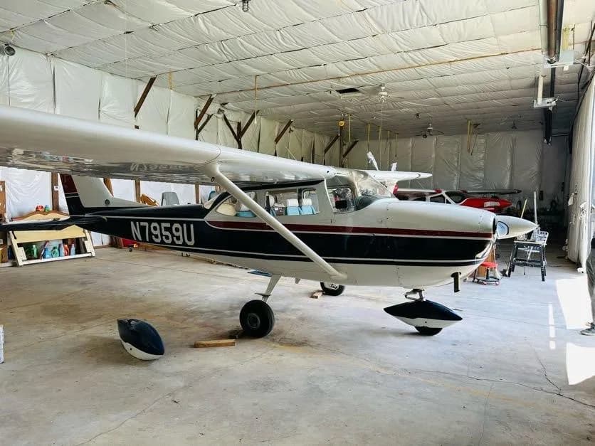 A three-quarter rear view of the white and blue Cessna N7959U with a red stripe in a hangar.