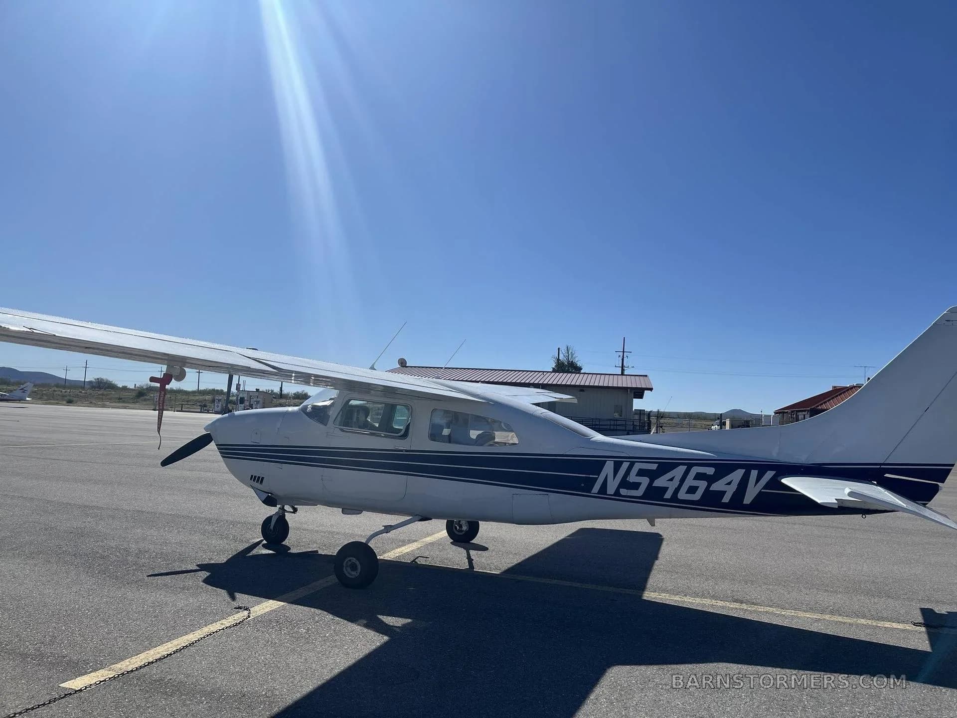 A white Cessna 182 with blue stripes, registration N5464V, parked on an asphalt ramp with the sun glaring.