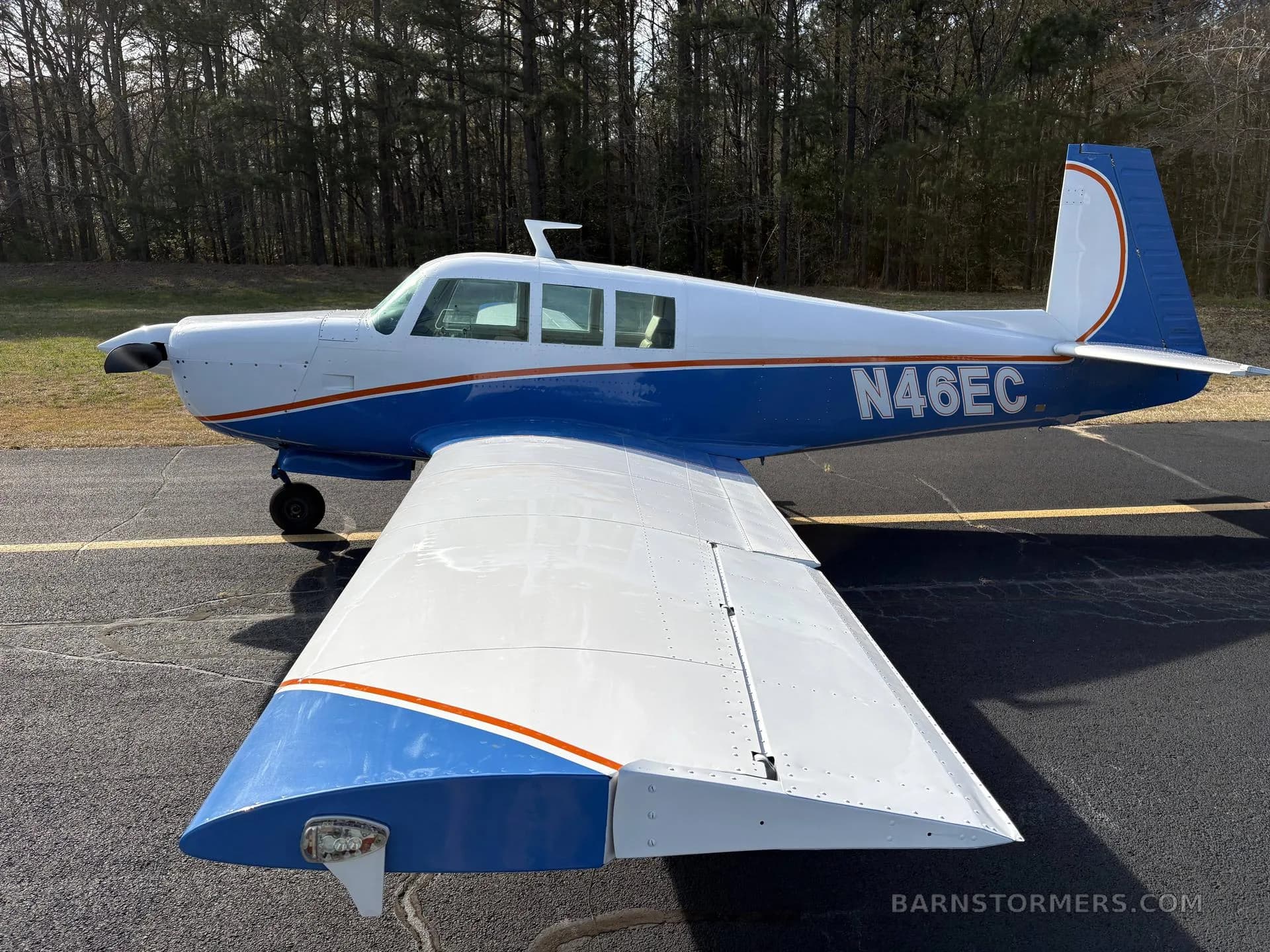 A white and blue single-engine aircraft with tail number N46EC parked on an asphalt ramp with a forest in the background.
