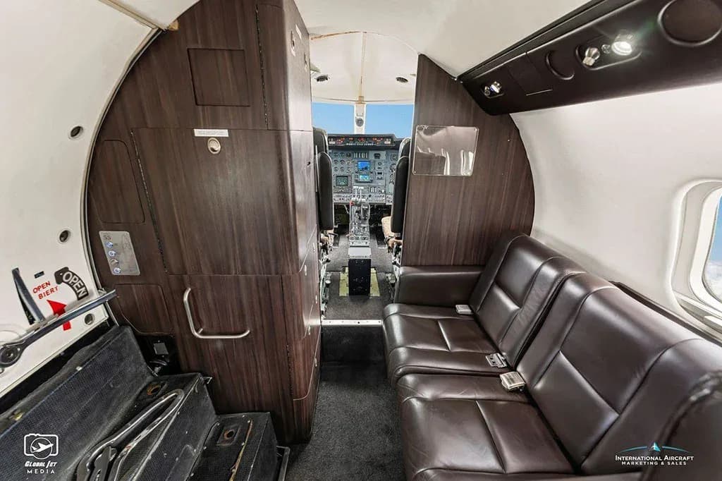 The interior cabin of a business jet with brown leather seating and a view into the cockpit through an open doorway.
