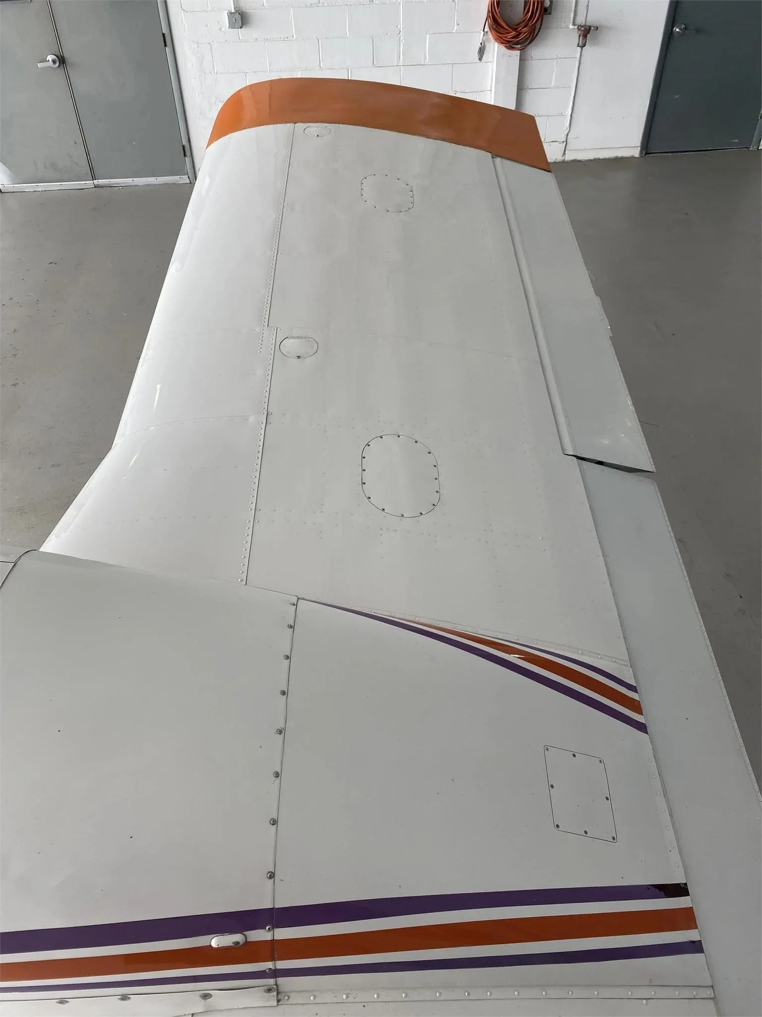 An overhead view of the left wing and fuselage of the white Piper Aztec aircraft, showing the orange wingtip and purple stripes.