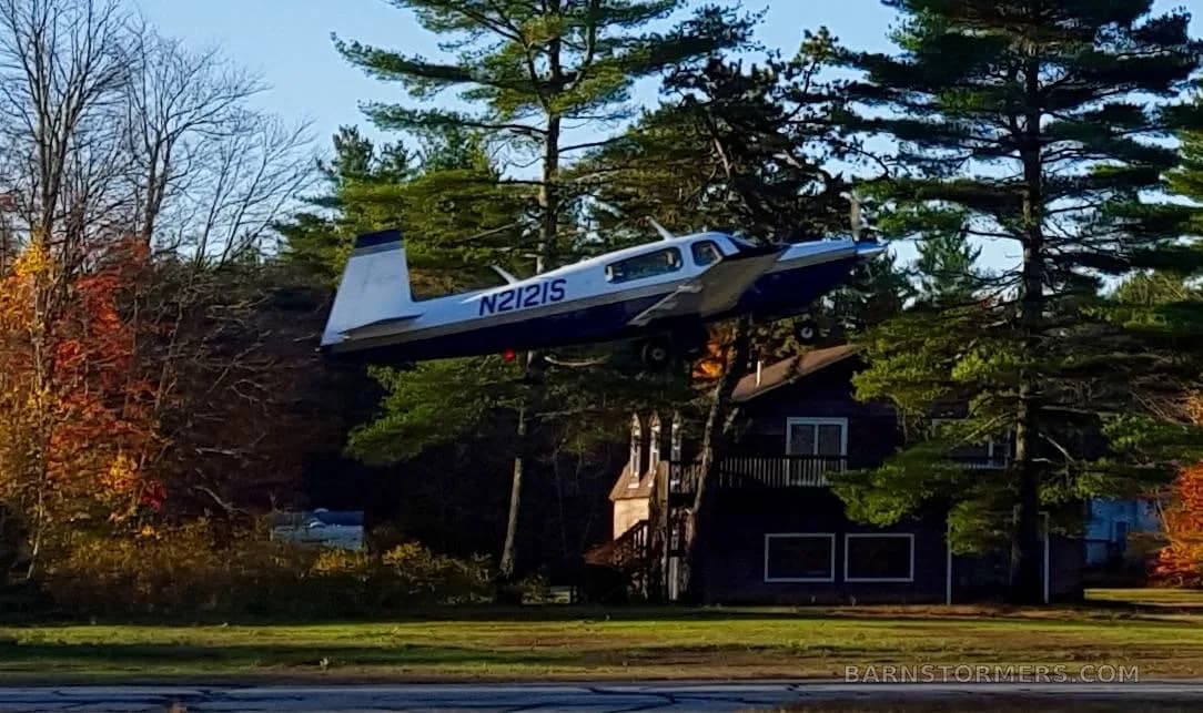 A white and blue single-engine propeller aircraft (N2121S) is seen flying low over a house and autumn trees.