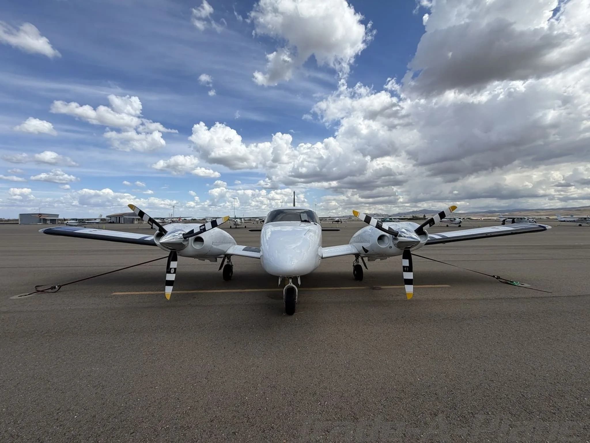 A head-on view of the white Piper Seneca II twin-engine aircraft with black and yellow striped propellers, tied down on an airport tarmac.