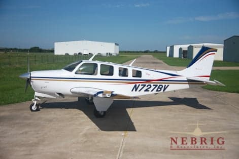 A white single-engine propeller aircraft, N727BV, with blue, gold, and red stripes, parked on a tarmac with hangars in the background.