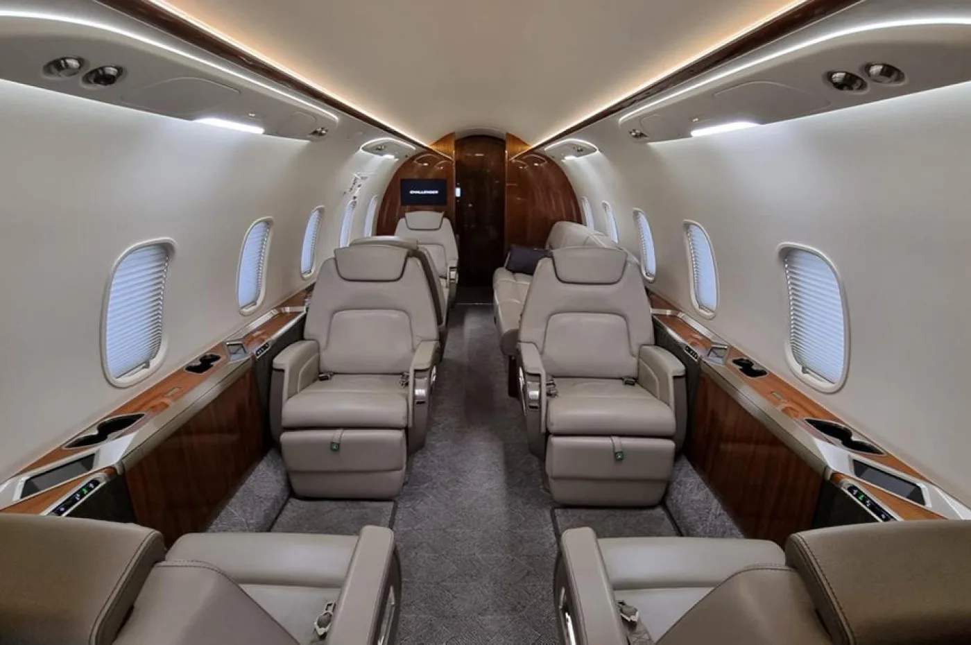 The luxurious interior cabin of a business jet shows beige leather seats arranged in a club configuration, with wood paneling and overhead lighting.