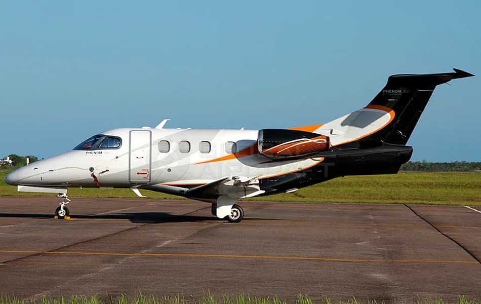 A white Embraer Phenom 100 business jet with black and orange livery parked on a tarmac under a clear sky.