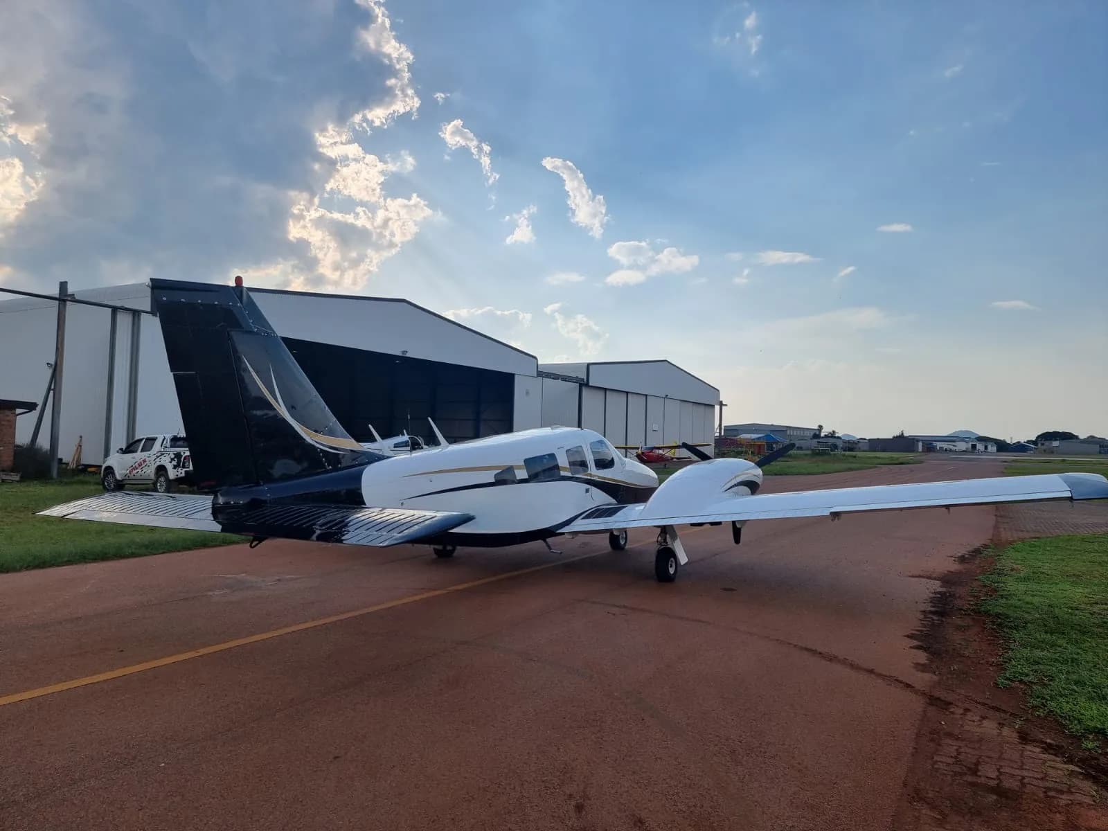 A white and black light twin-engine aircraft with gold accents is parked on a tarmac in front of hangars under a partly cloudy sky.