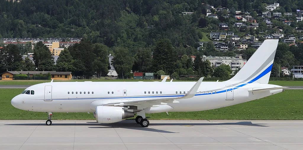 A white Airbus A320 family aircraft with blue and grey stripes on the tail and fuselage, parked on an tarmac with a green hillside and town in the background.