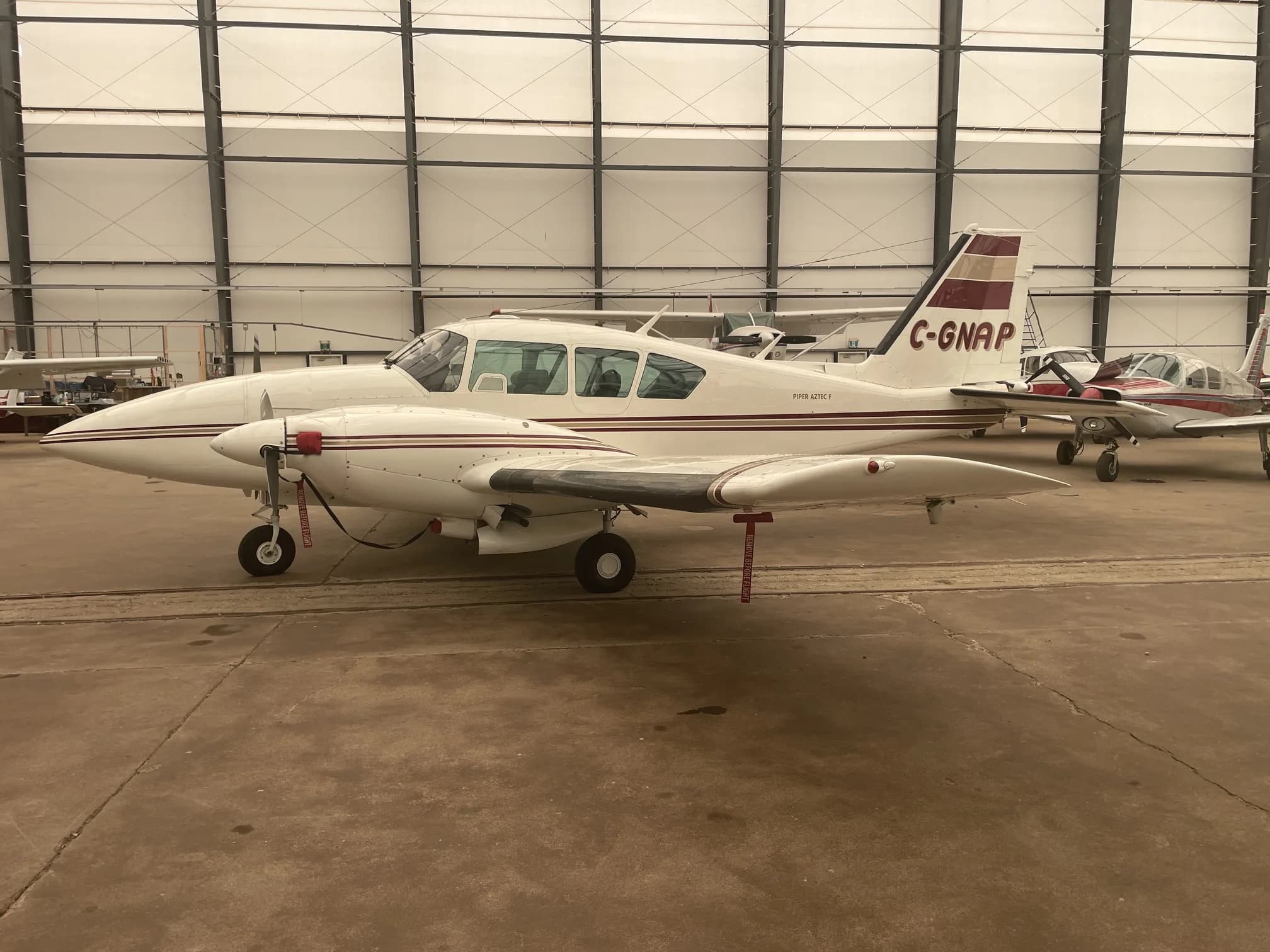 A white and maroon twin-engine Piper Aztec aircraft, registration C-GNAP, parked inside a hangar.
