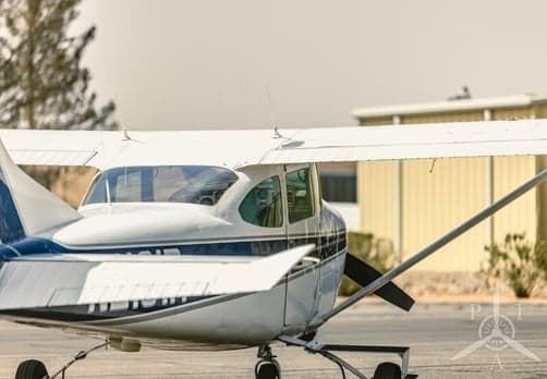 A rear-quarter view of a white Cessna 182 with blue stripes parked on an airfield, showing the tail and wing.