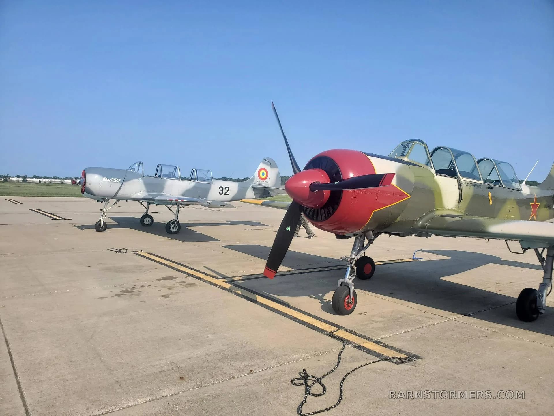Two Yakovlev Yak-52 aircraft parked on an airfield, one in green camouflage with red stars and another in grey with a Romanian flag on the tail.