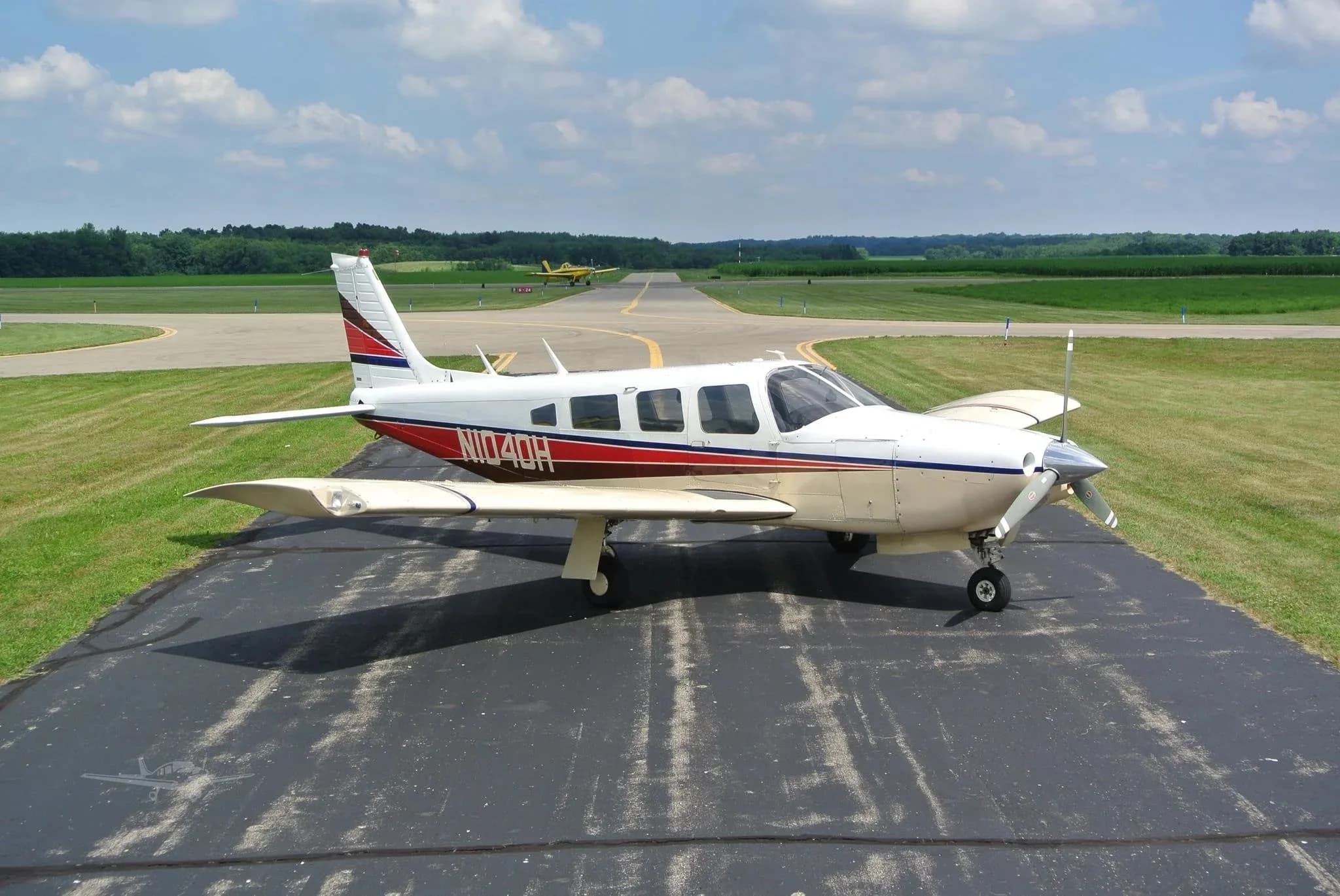 A white and beige single-engine aircraft with red and blue stripes parked on an asphalt runway with another aircraft in the distance.
