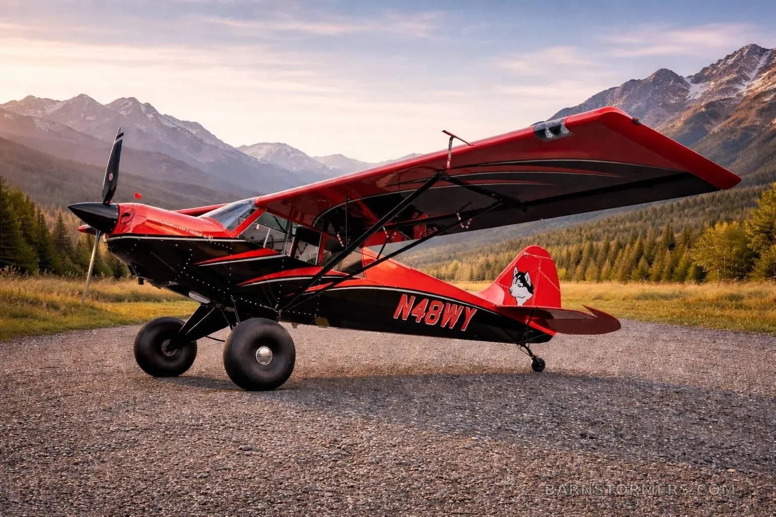A red and black Aviat Husky A-1C aircraft with large tundra tires is parked on a gravel path with mountains and trees in the background.