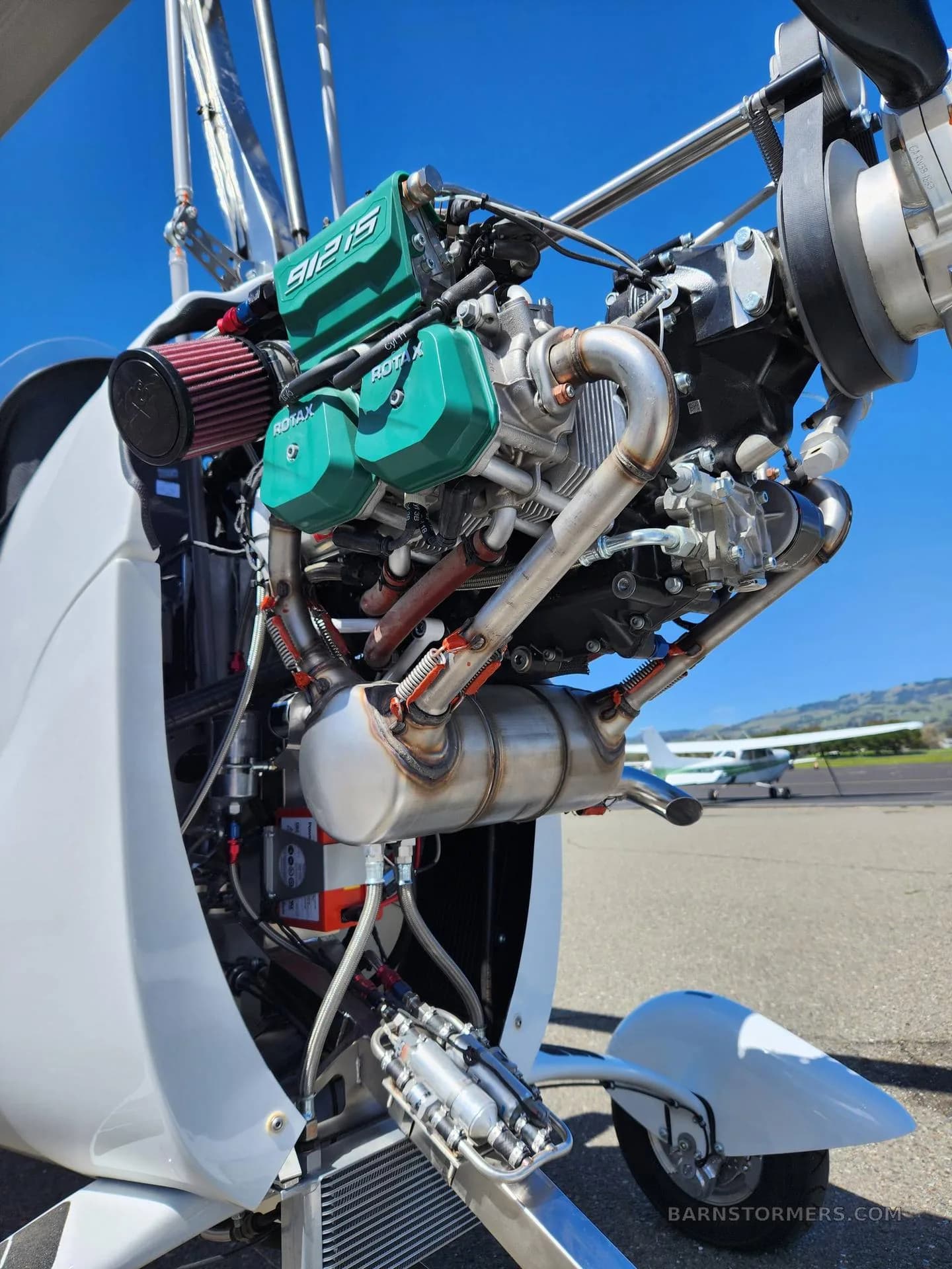A detailed view of the exposed Rotax 912iS engine of a white gyrocopter, showing its components and exhaust system.