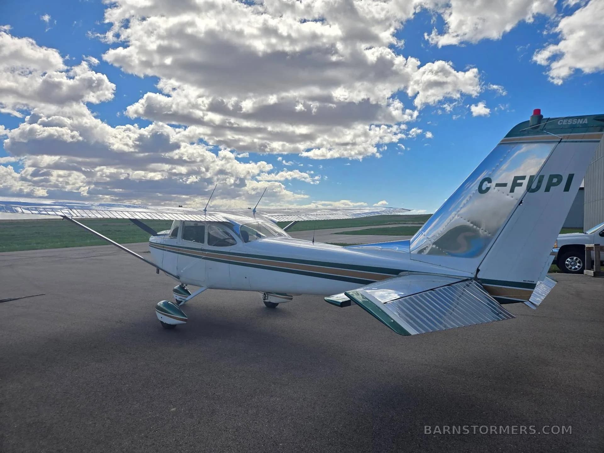 A white and green Cessna 172 with registration C-FUPI is parked on the tarmac under a partly cloudy blue sky.