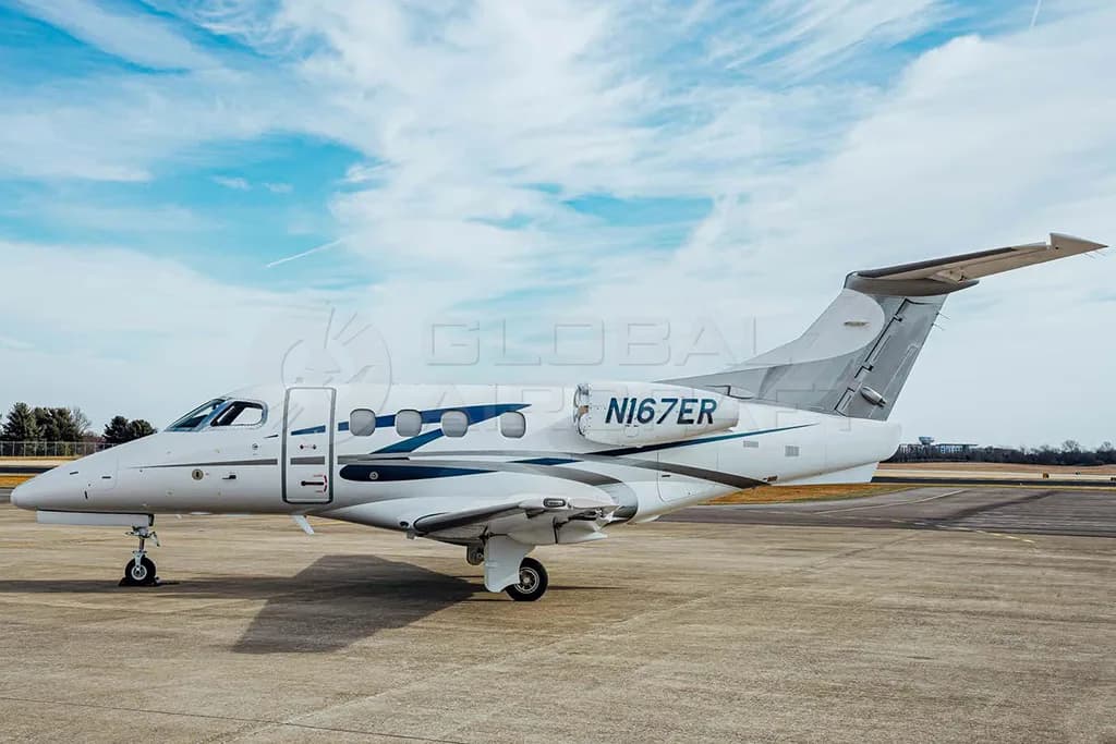A side view of a white Embraer Phenom 300 business jet with blue and grey stripes, parked on an airfield.