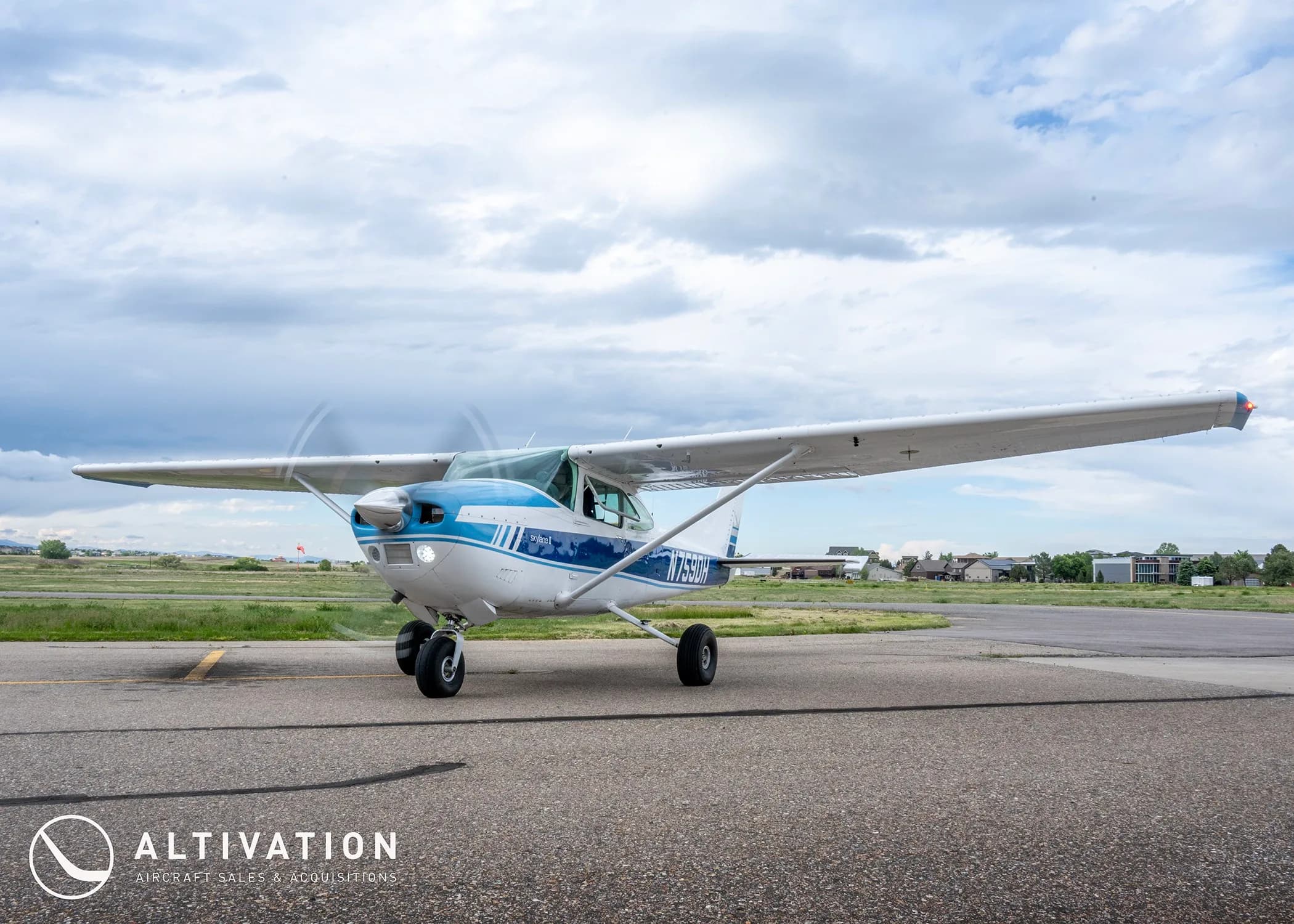 A white and blue Cessna 182 Skylane II (N759DH) with its propeller spinning, on an airfield under a cloudy sky.