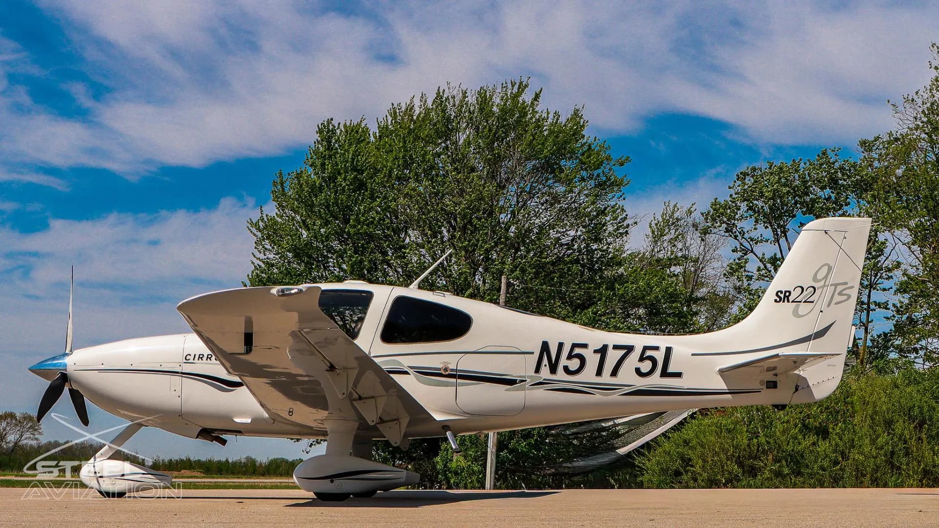 Left rear three-quarter view of a white Cirrus SR22 aircraft (N5175L) with its tail and wing visible, against a blue sky.