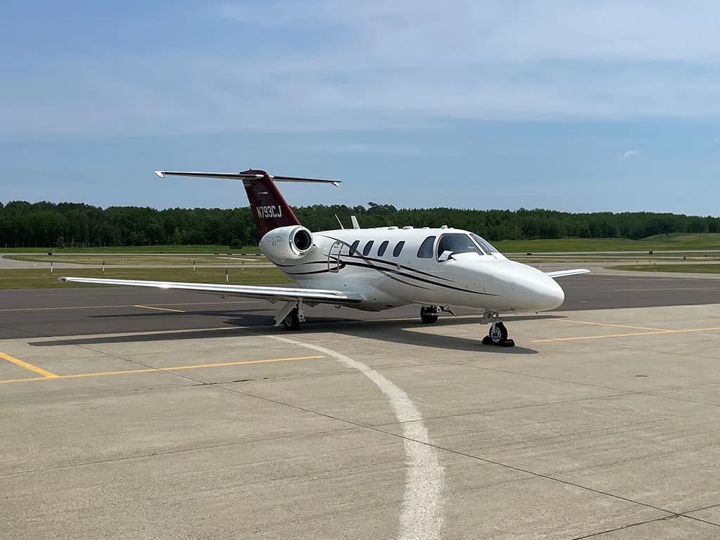 A white Cessna Citation CJ1 private jet with red and blue stripes, parked on an airport runway under a clear sky.