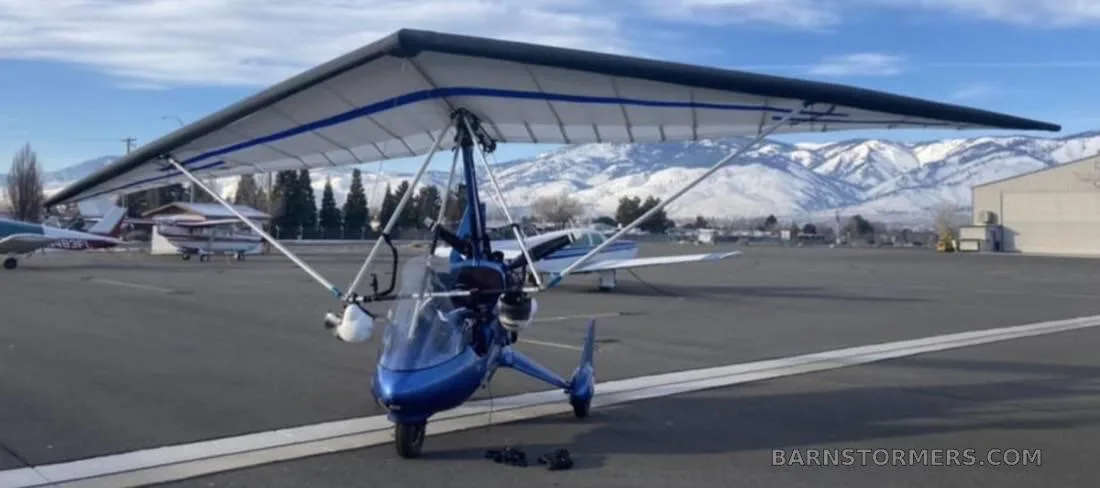 A blue ultralight aircraft with a white and blue wing is parked on an asphalt runway with snow-capped mountains in the background.