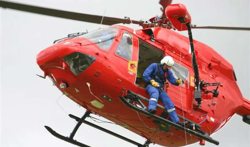 A red helicopter in flight with a person in a blue suit and white helmet standing on its skid.