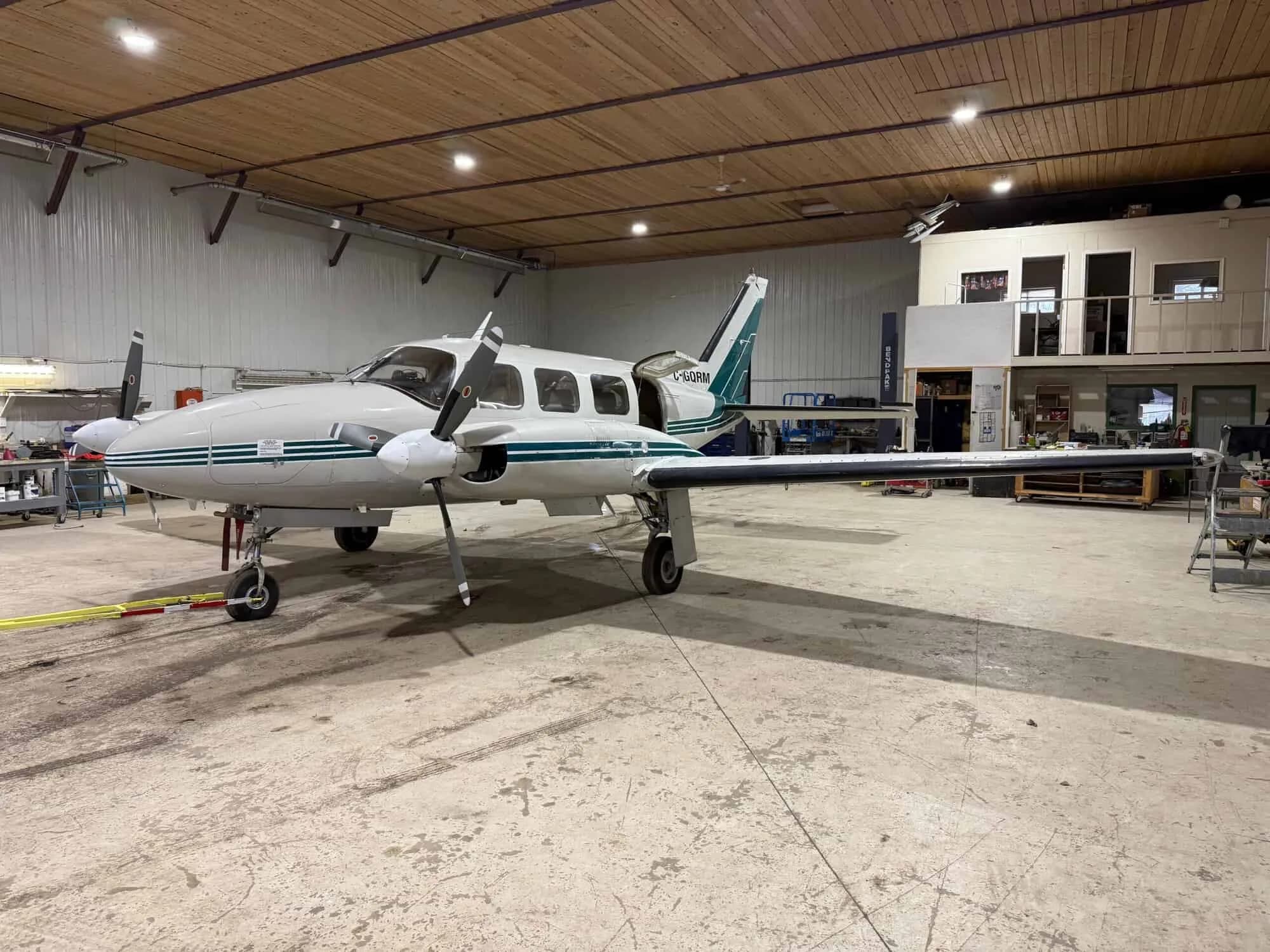A white and green twin-engine aircraft, registration C-GQRM, parked in a hangar with its nose facing right.