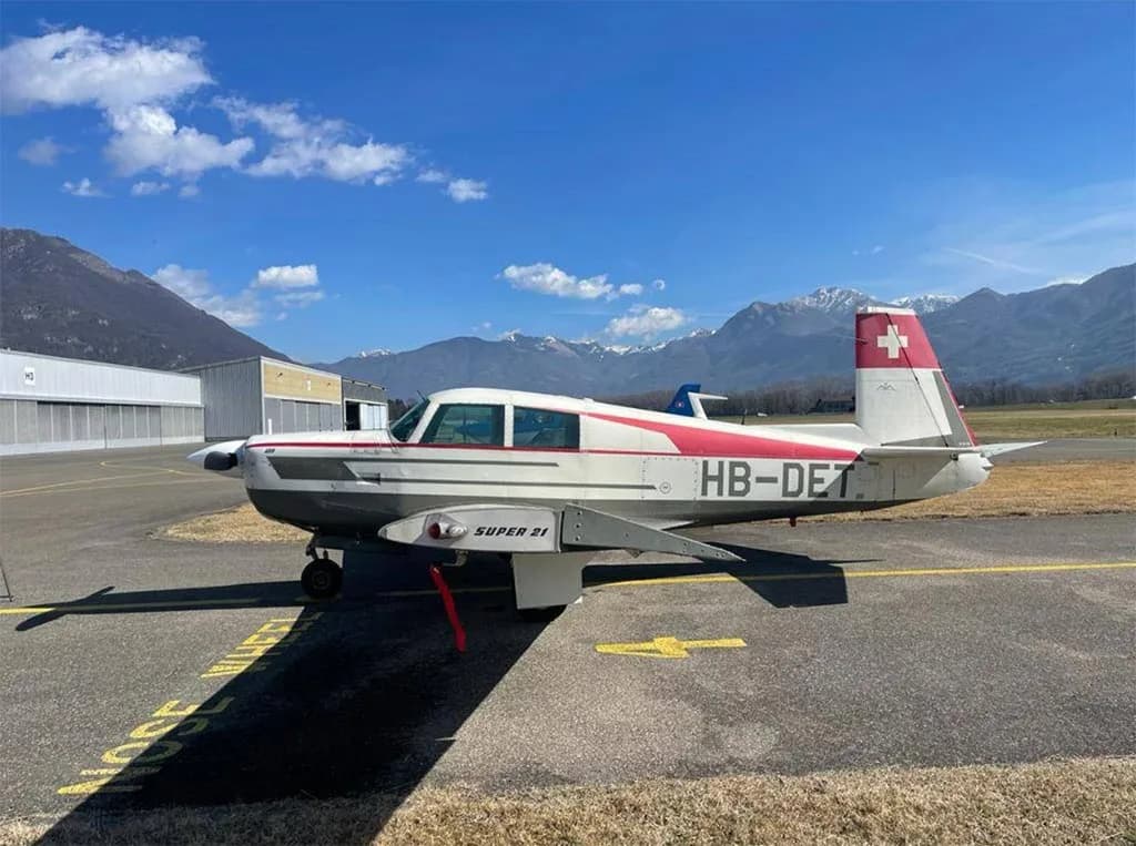 A white and red single-engine aircraft, HB-DET, with a Swiss flag on its tail, parked on an airfield with mountains in the background.