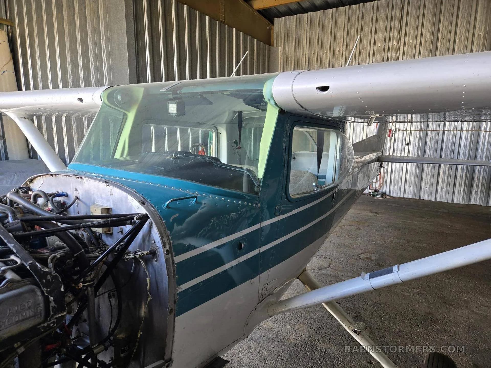 A close-up view of the front of a light aircraft in a hangar, with the engine cowling removed, showing the engine and propeller shaft.