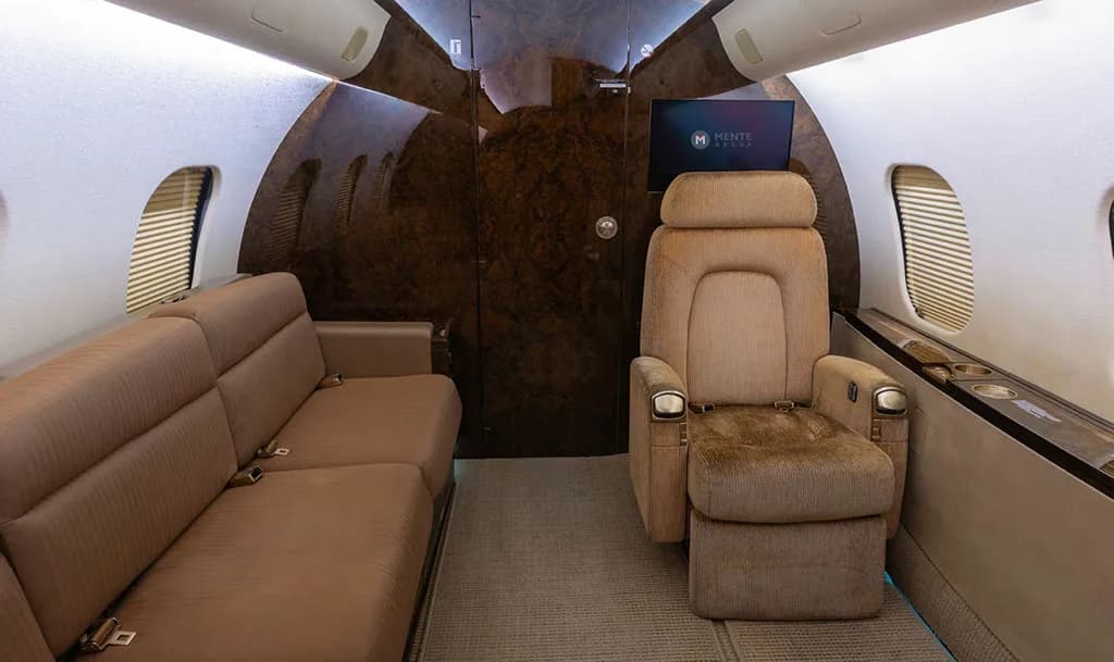 The luxurious interior cabin of a private jet, featuring a tan sofa, an individual plush tan seat, and dark wood paneling.