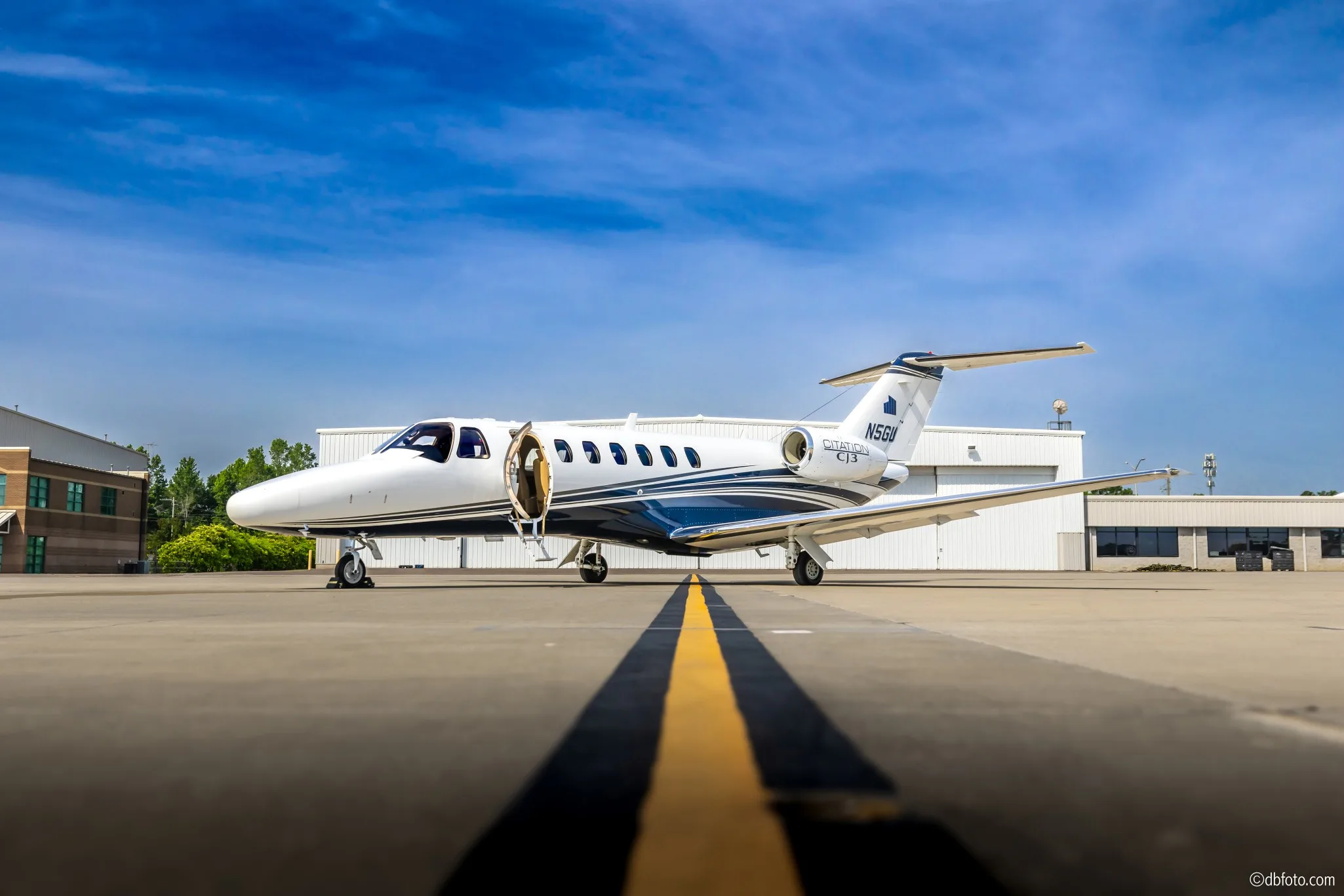 A white Cessna Citation CJ3 business jet with blue and grey stripes, registration N5GU, with its main door open, parked on an airport tarmac.
