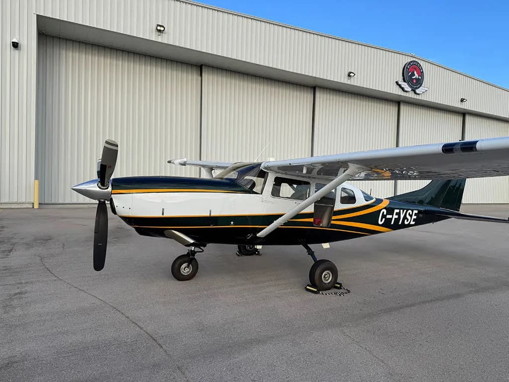 A dark green and white Cessna 182 with yellow stripes, registration C-FYSE, parked on an airfield in front of a hangar.