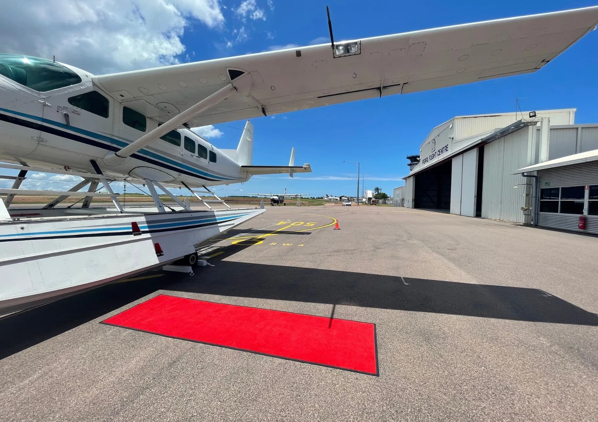A seaplane parked on the tarmac with a red carpet leading to the door and a hangar in the background.