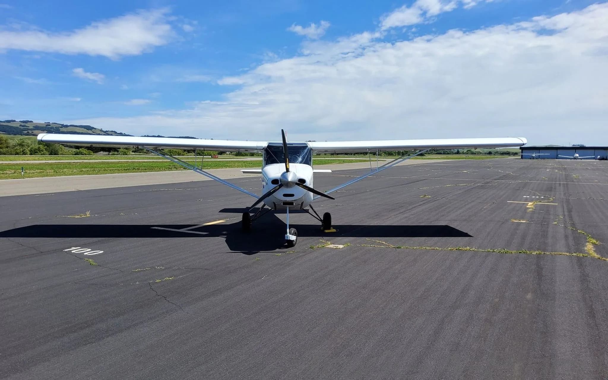 A white BushCaddy aircraft with a propeller, viewed from the front on a sunny tarmac with hills in the background.