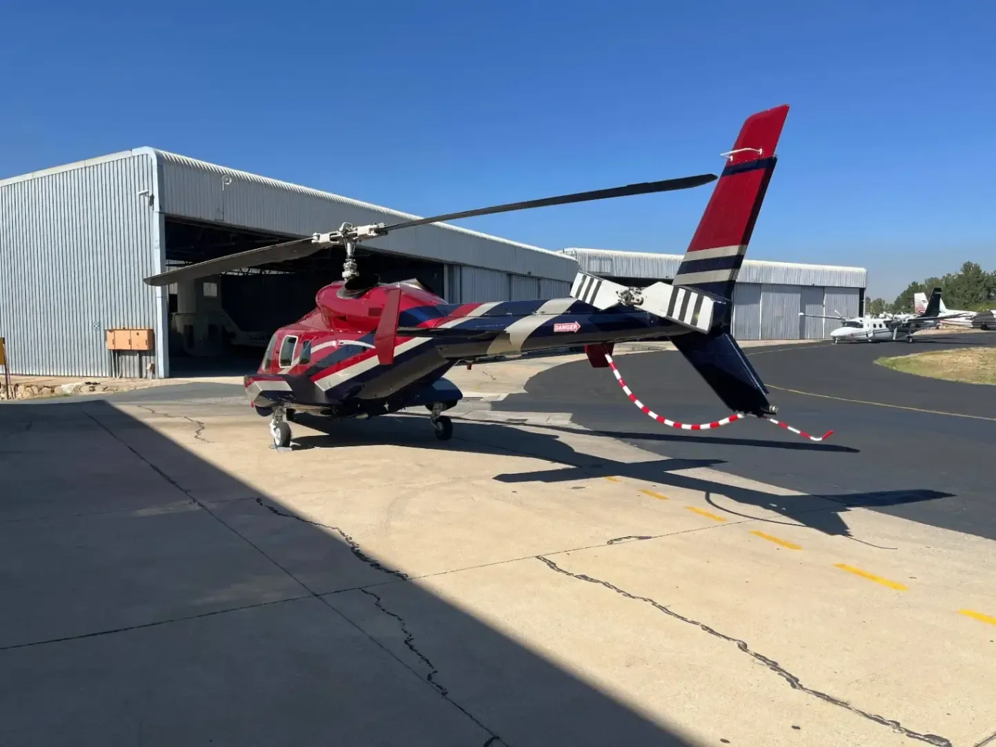 A red and white helicopter sits on the tarmac in front of a hangar.