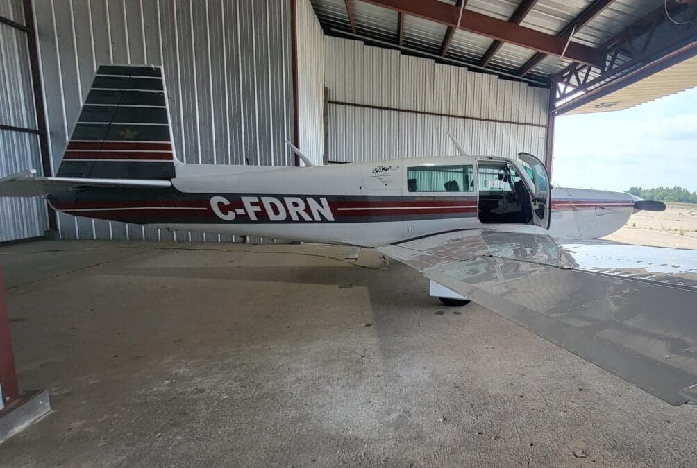 A white Mooney M20J aircraft with red and grey stripes, registration C-FDRN, parked inside a hangar with its door open.