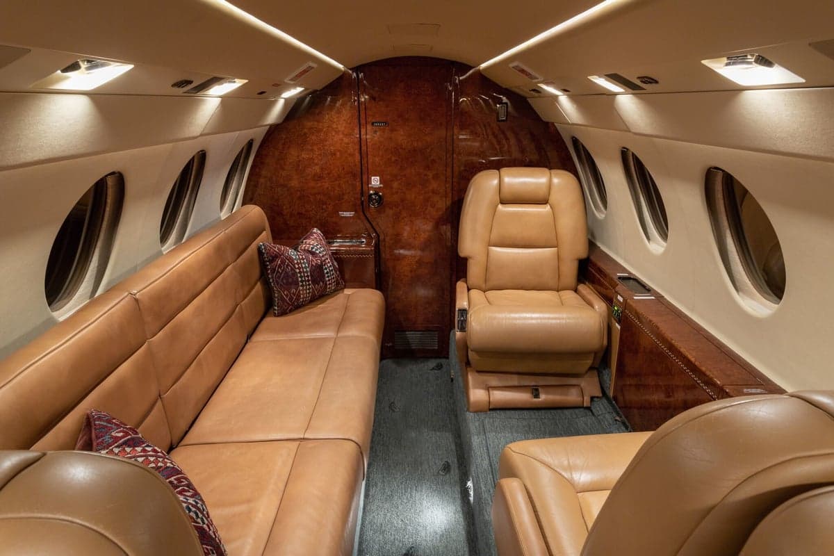 A spacious private jet cabin with a long tan leather sofa, individual seats, and elegant wood paneling.
