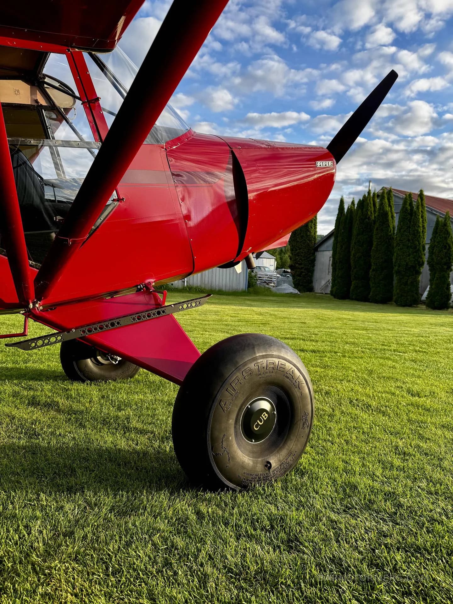 A close-up view of the front propeller, engine cowling, and large "CUB" tire of a red Piper Cub aircraft on green grass.