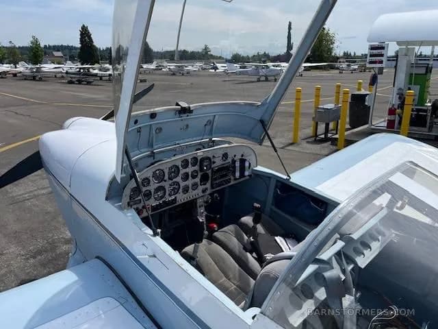The open canopy and cockpit of a light blue and white aircraft, showing the instrument panel and seats, with an airfield in the background.
