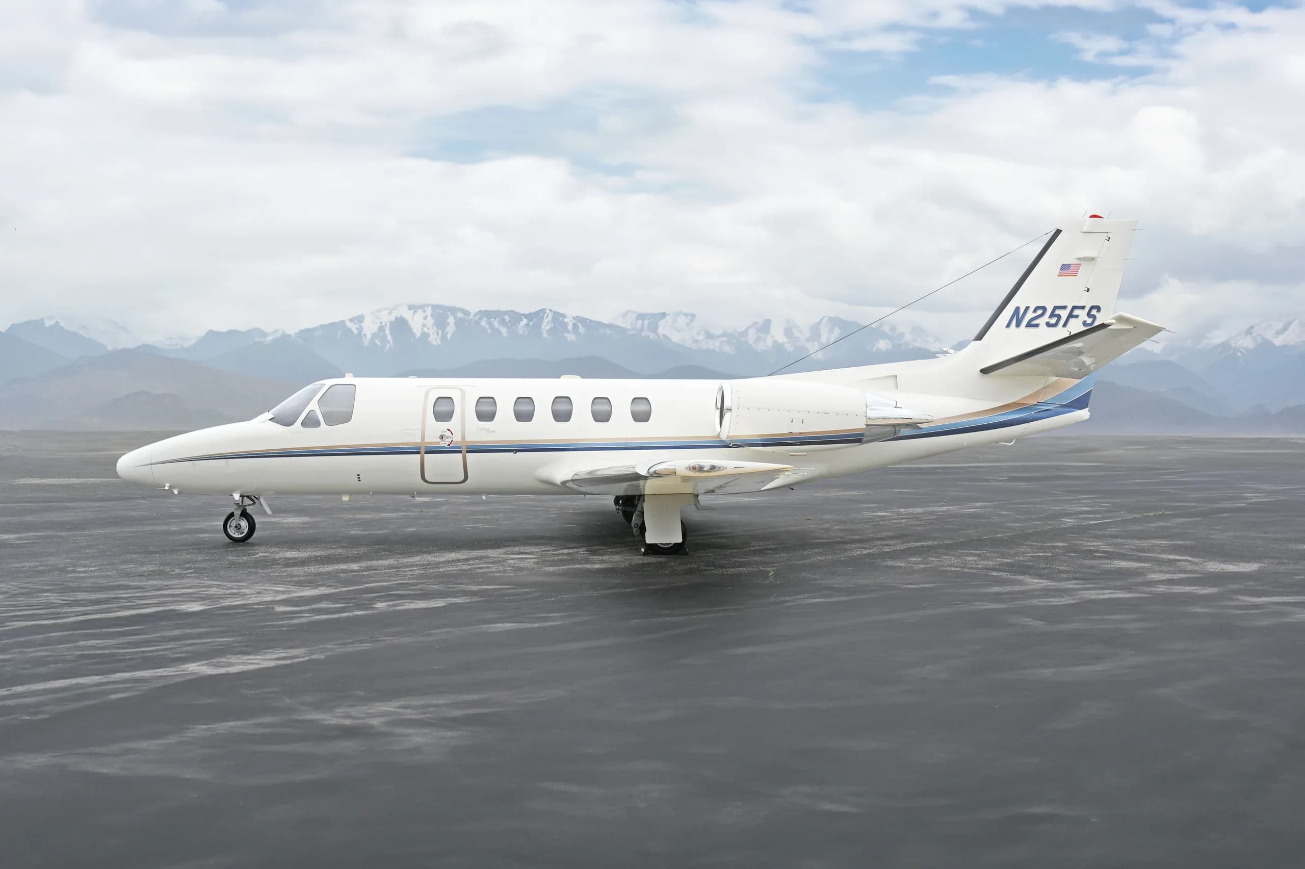A side view of a white Cessna Citation business jet (N25FS) with blue and gold stripes, parked on an asphalt surface with snow-capped mountains in the background.