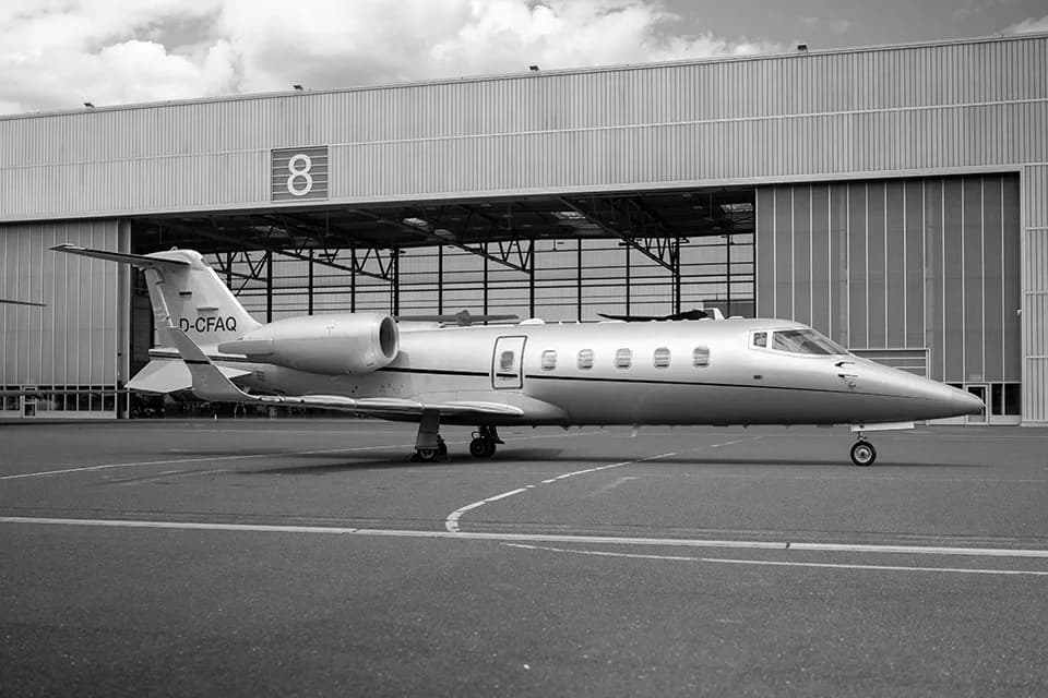 A silver business jet, registration D-CFAQ, parked on an airfield in front of a large hangar.