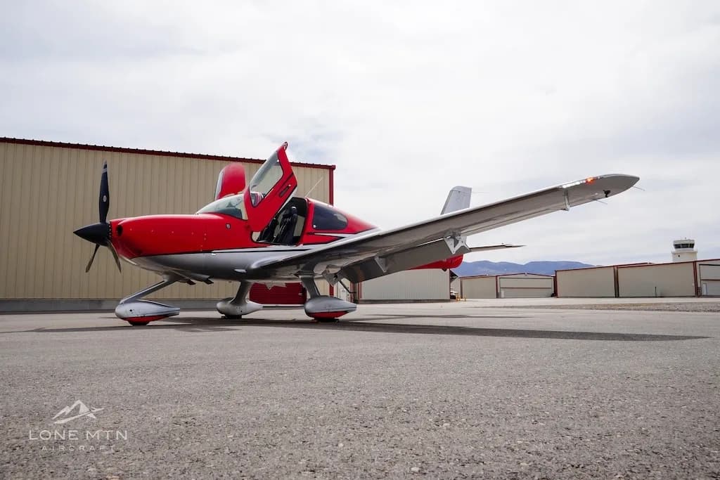 A low-angle front-side view of a red and silver Cirrus SR22 aircraft with open doors, parked on an airfield with a control tower in the distance.