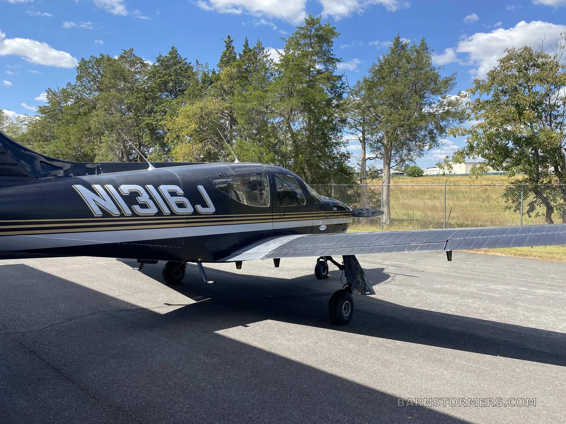 A black Cessna 112A aircraft with gold and silver stripes, registration N316J, parked on asphalt under a partly cloudy sky.
