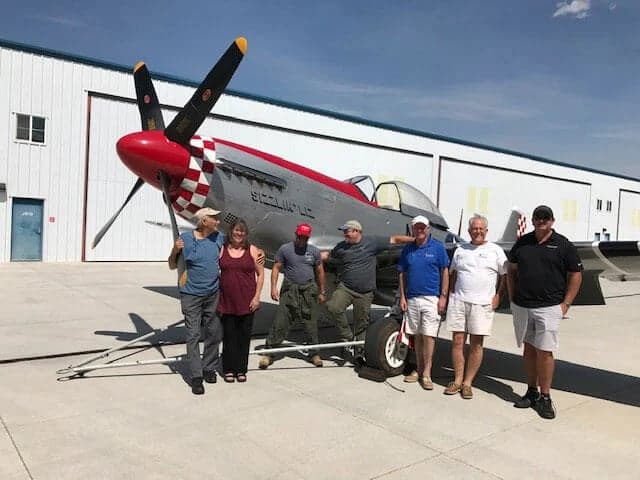 A silver P-51 Mustang with a red nose and checkerboard pattern, parked on an tarmac with a group of people.