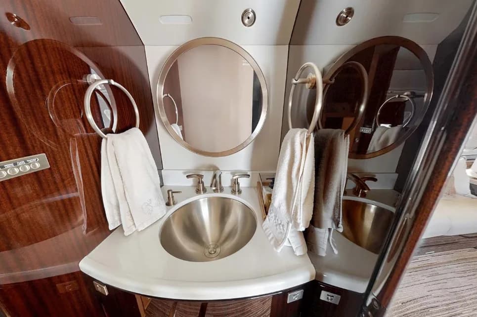 The aircraft lavatory features a polished metal sink, a round mirror, and dark wood paneling.